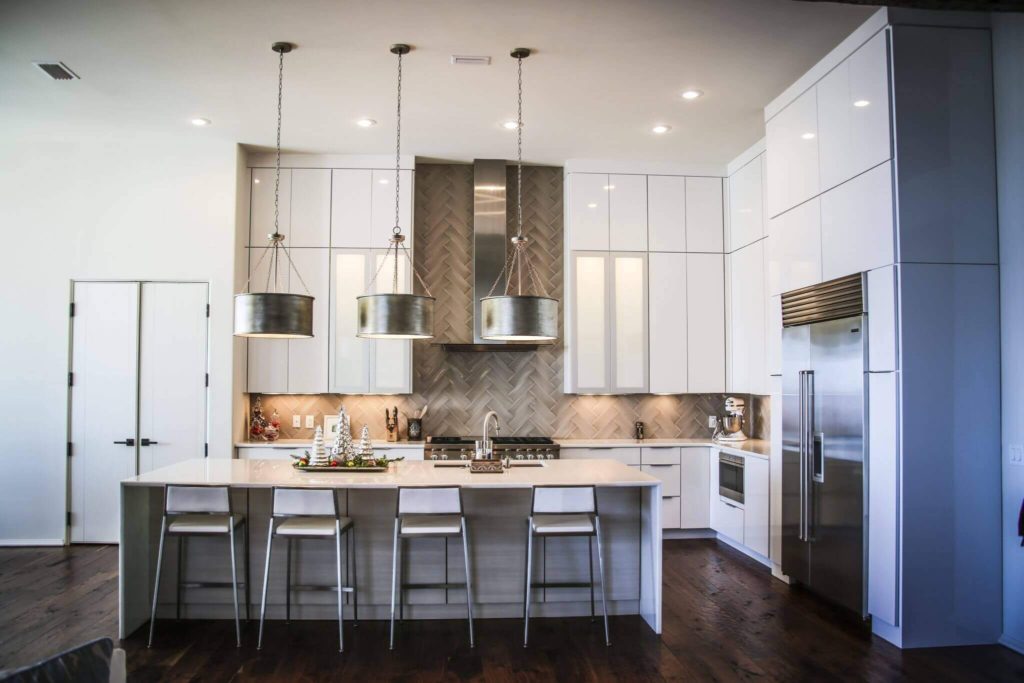 Modern kitchen with high ceilings, white, sleek cabinetry, an island with white stools, and large hanging lights over the island.