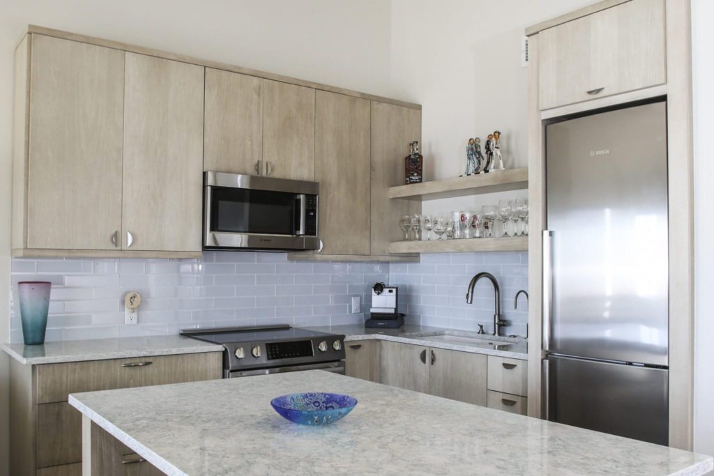 Costal inspired kitchen with light wood cabinets, stainless steel fridge and microwave, and marble countertops.