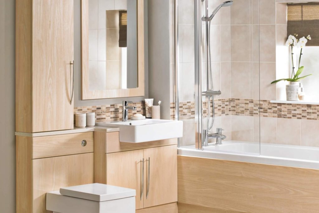 Light wood coloured bathroom with built-in cabinets, a small vanity with a white sink, and a tub with glass doors and a shower faucet.