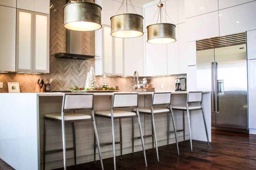 Contemporary Kitchen with white cabinetry and tall, white chairs at an island. Large modern lights hang over the island and is a stainless steel fridge.