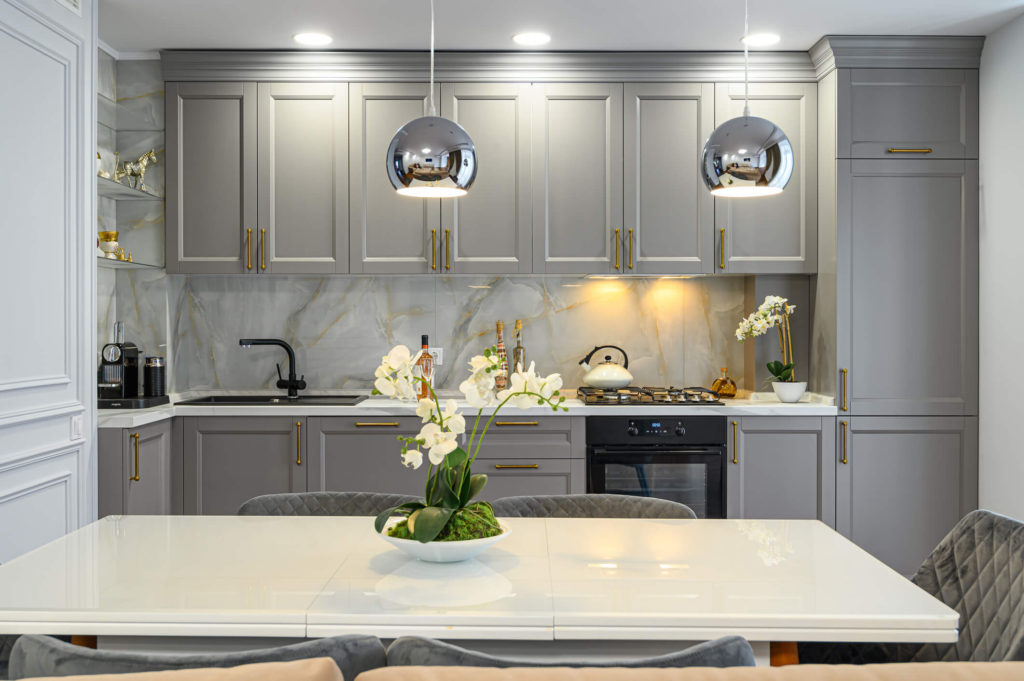 Contemporary kitchen with grey Shaker cabinets, a white table with grey chairs and flowers on the table, and 2 silver hanging lights.