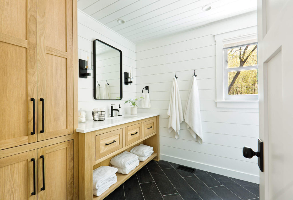 Coastal inspired bathroom with white shiplap walls, dark tile floors, and custom wooden cabinetry with a white countertop and sink.