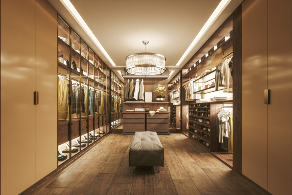 Long, modern, walk-in closet with custom built-ins, a fancy chandelier, and a bench in the centre. Closets are filled with clothing.