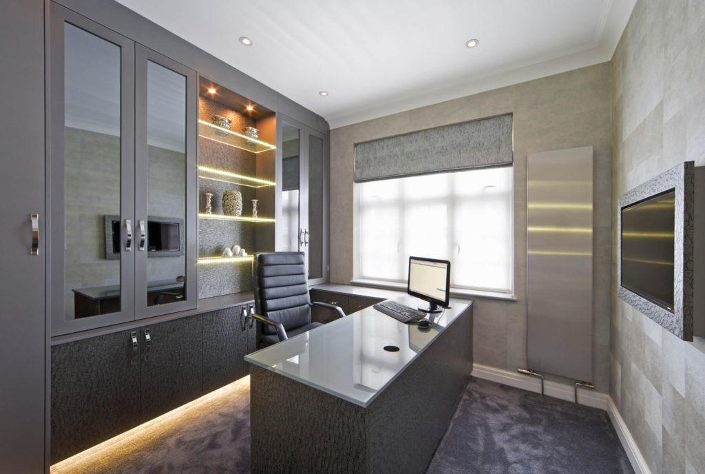 Office with built-in storage cabinets, glass doors, open shelving, and lighting in the cabinet. A chair and desk are centered in the room in front of the shelving.