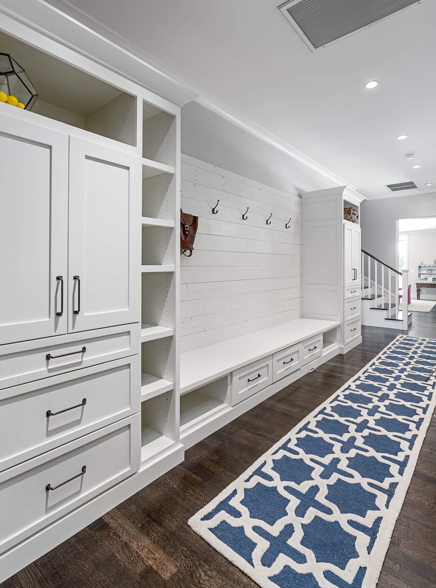 Mudroom/hallway/entryway with white built-in cabinets, low storage drawers, and a bench. All in white wood. There is a long blue and white runner on the dark wood floor.