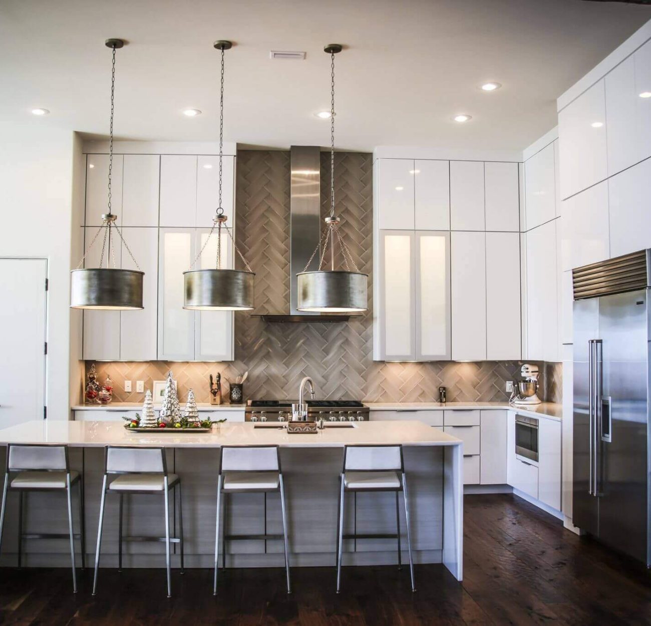 Modern kitchen with high ceilings, white, sleek cabinetry, an island with white stools, and large hanging lights over the island.